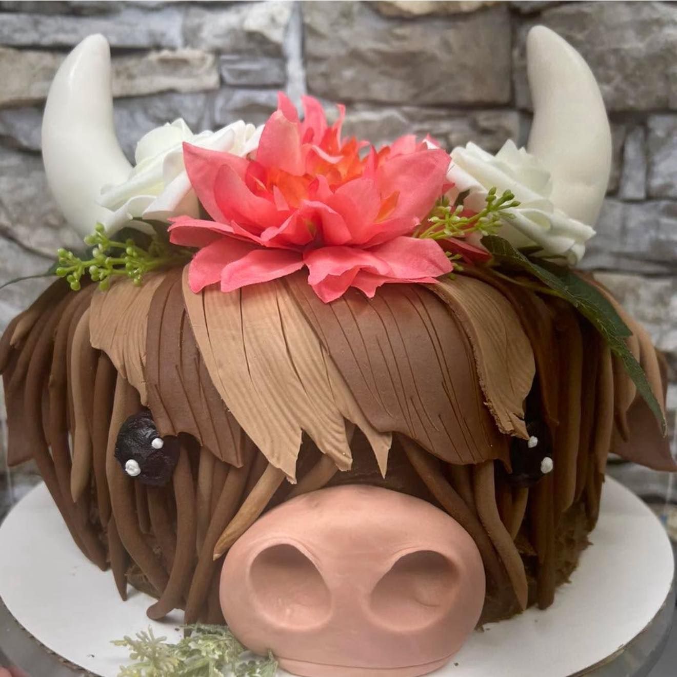 Cake shaped like a highland cow with floral crown and horns. Brown and pink details.