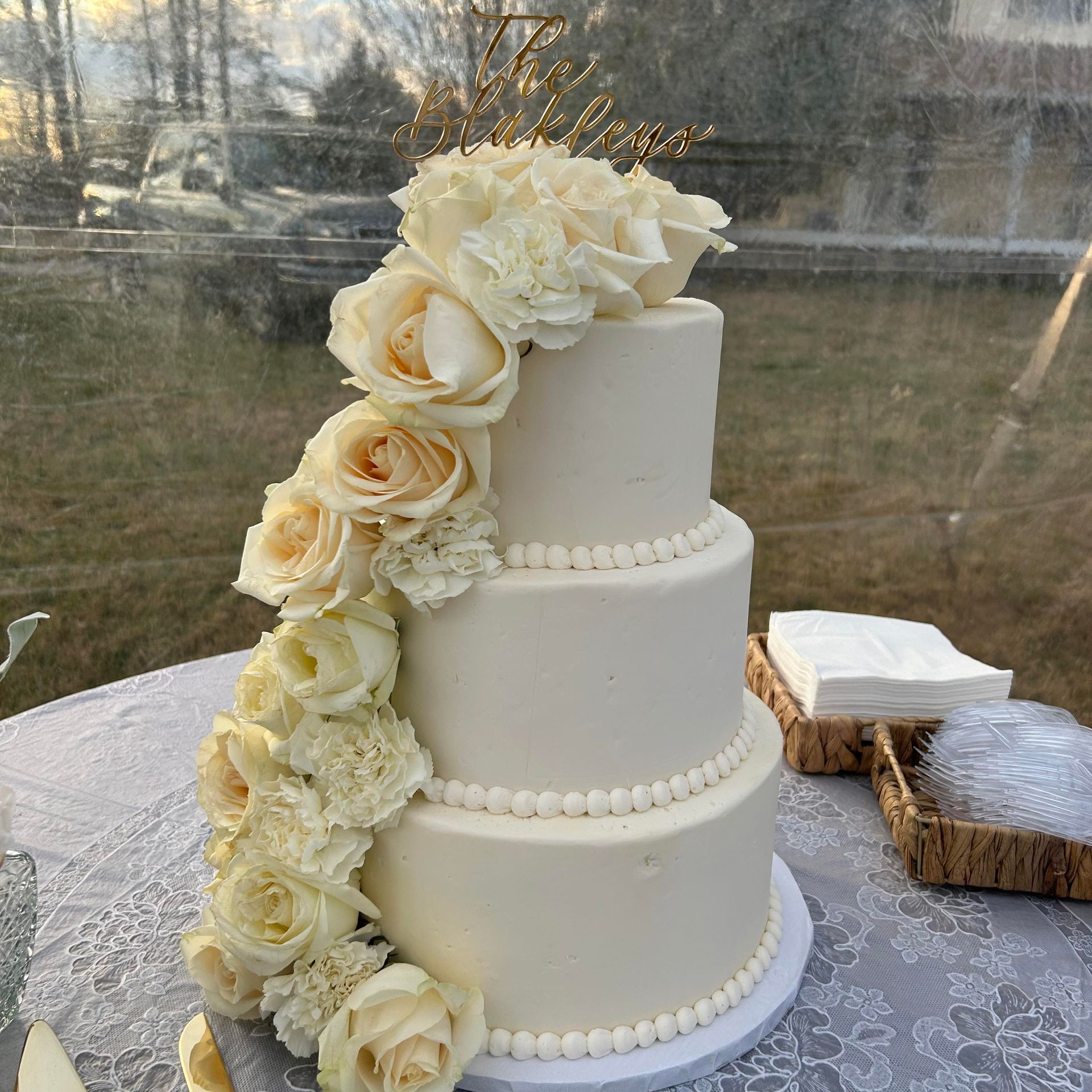 Three-tiered white wedding cake with cascading light roses on a white table with a garden backdrop.