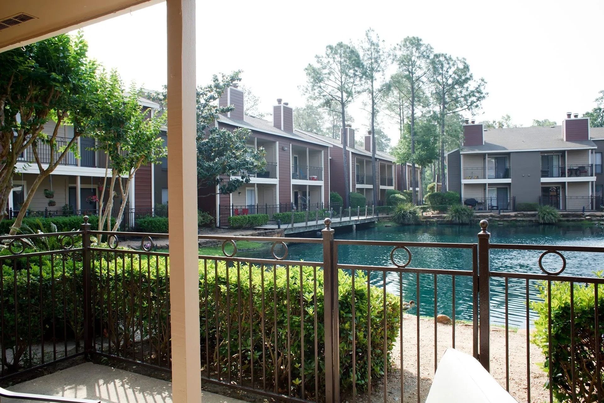 View of a lakeside apartment community with two-story buildings, balconies, and a metal railing along a pond.