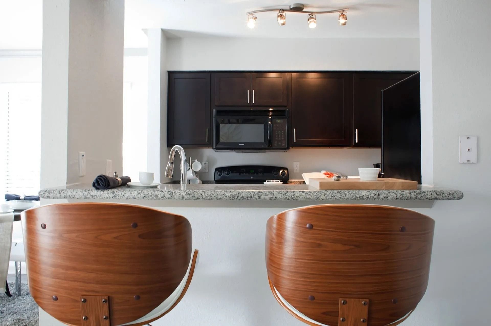 Kitchen with granite counter, dark wood cabinets, stainless microwave and stove, and wooden bar stools.