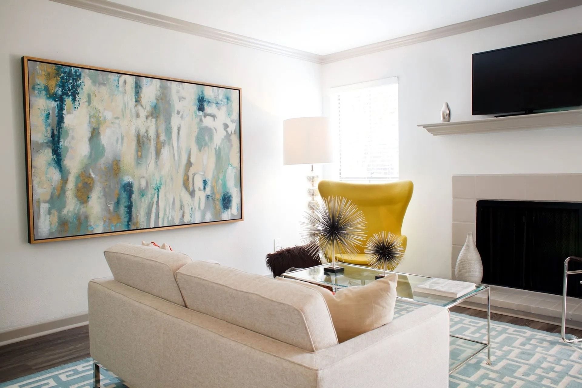 Bright apartment living room with beige sofa, yellow accent chair, glass coffee table, and abstract wall art.