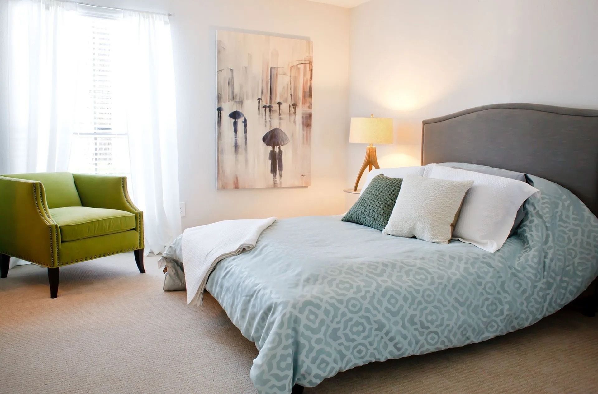 Bedroom in an apartment with a bed, blue patterned duvet, green accent chair by the window, and a lamp.