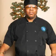 Man in black chef coat and hat standing indoors, with a decorative tree behind him.