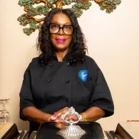 Chef standing behind a table, wearing a black jacket and glasses, with hands resting on a decorative metal object.