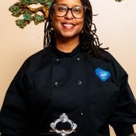 Person smiling in a black chef coat with a blue logo, standing indoors against a beige wall.