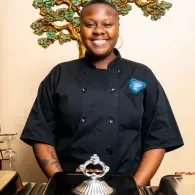 Chef in a black uniform smiling behind a counter with a decorative wall piece behind her
