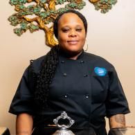 Chef in a black uniform standing indoors, with decorative wall art behind her.