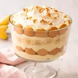 Layered dessert in a glass bowl, topped with whipped cream and chopped nuts, with bananas nearby