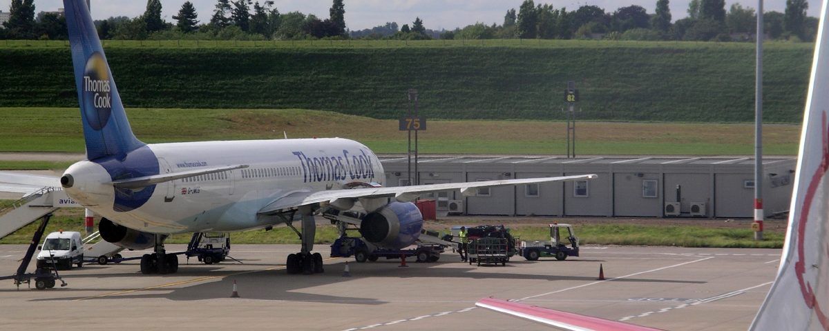 An airplane with a blue tail and white body parked at an airport. Green field and buildings in background.