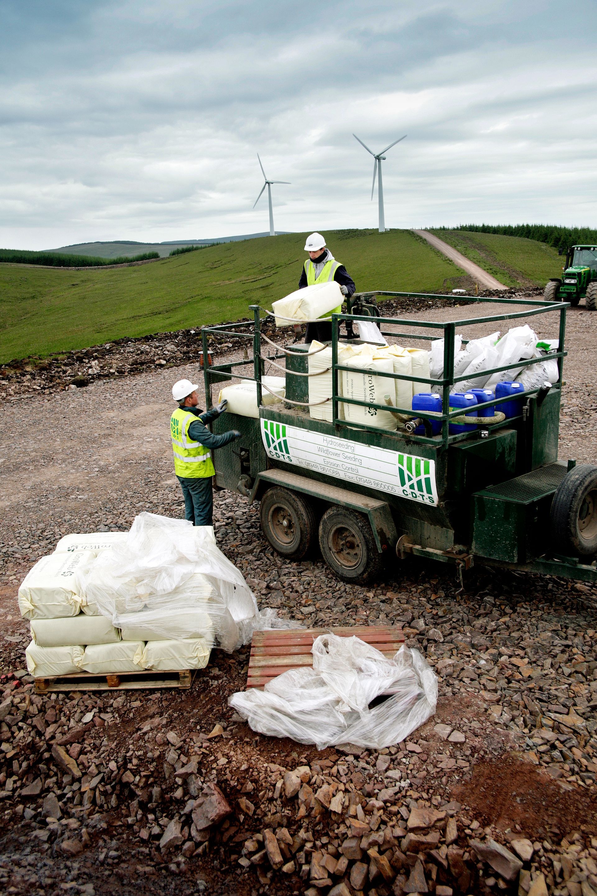 Two workers loading materials into a trailer at a construction site near wind turbines.