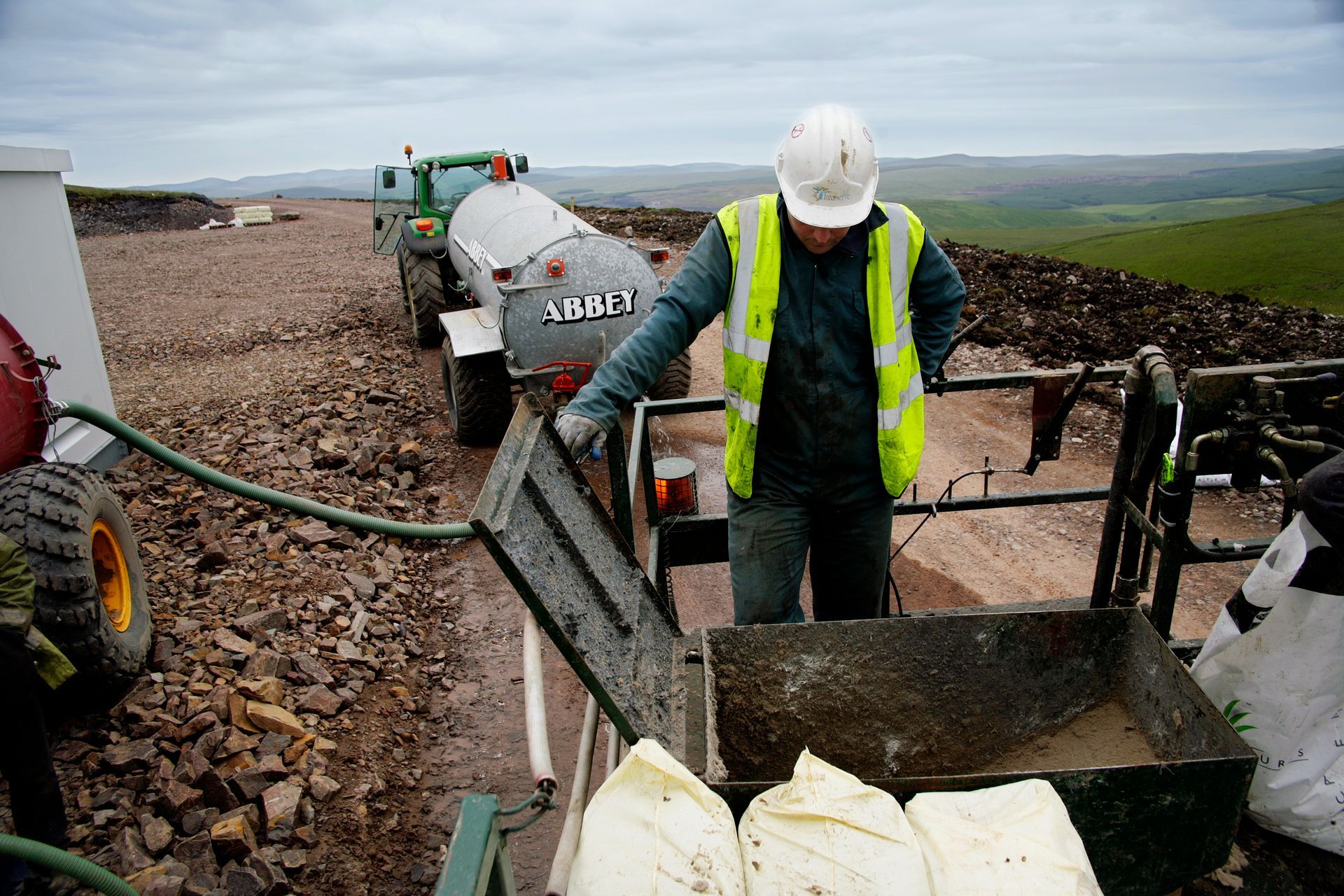 Worker in hard hat and safety vest opening a tank lid, with a truck and terrain in the background.