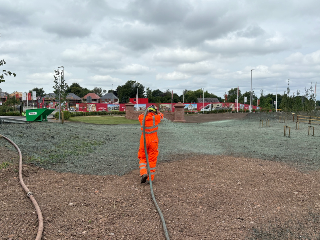 Person in orange suit sprays a large field with a hose, construction site in the background under a cloudy sky.