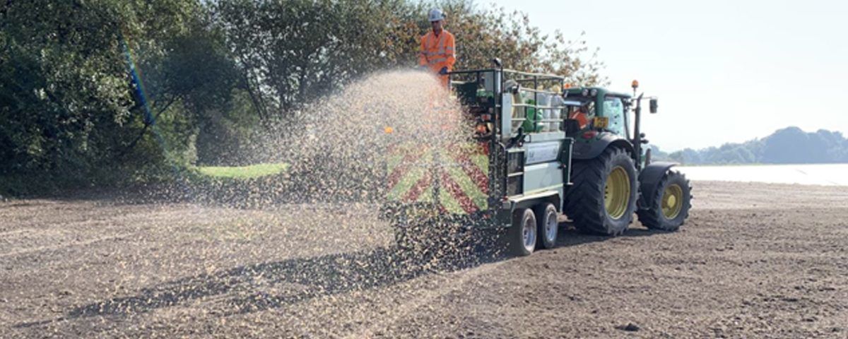 A tractor spreads material on a sandy field near a body of water and treeline. A person is visible on the tractor.