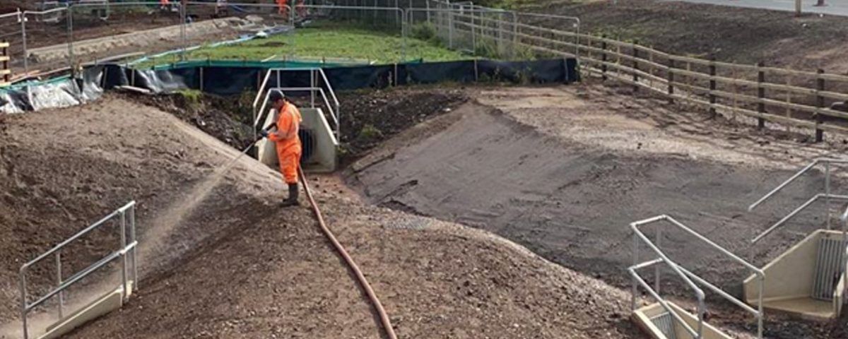 A worker in orange overalls sprays water on a gravel embankment at a construction site.