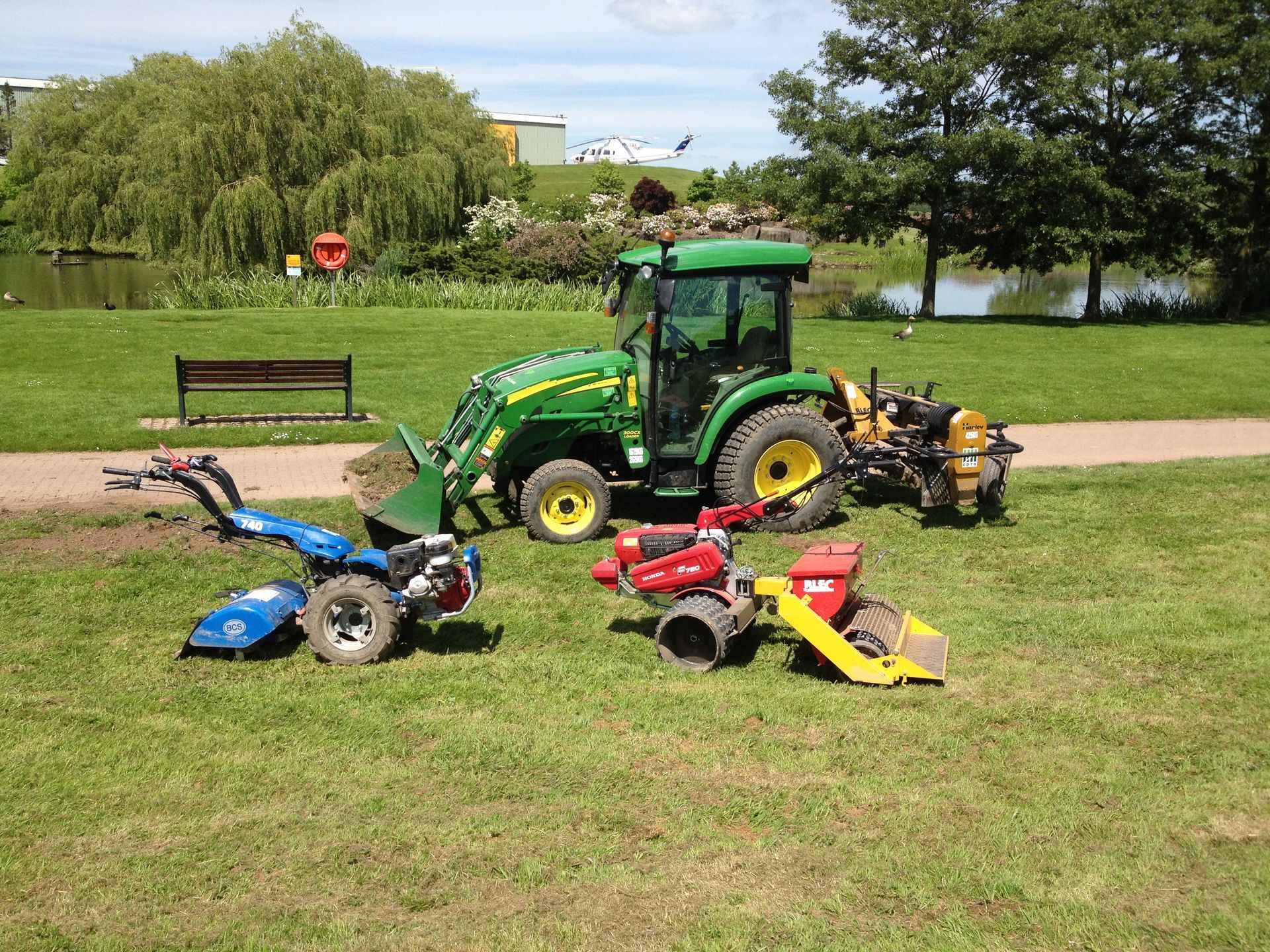 Several green and red tractors and tillers on a grassy area near a lake.