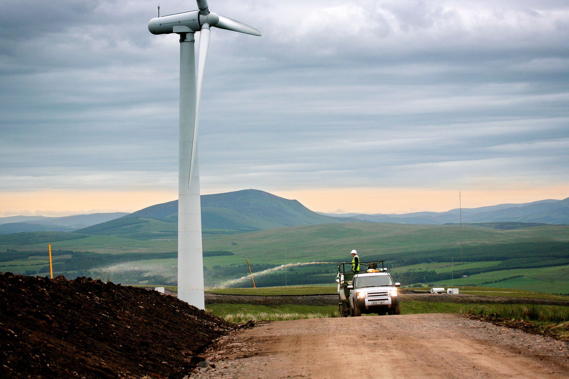 Wind turbine on a hill with a white truck; workers in the distance. Overcast sky, green landscape.