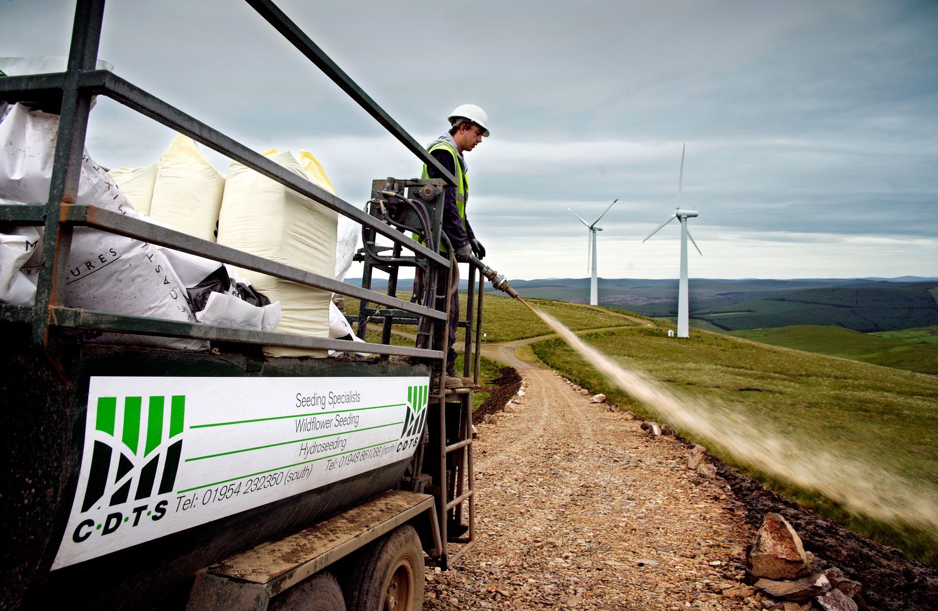CDTS operative loading hydroseeding equipment on a lorry-mounted unit at Clyde Wind Farm, spraying seed and mulch mix onto disturbed ground alongside an access track