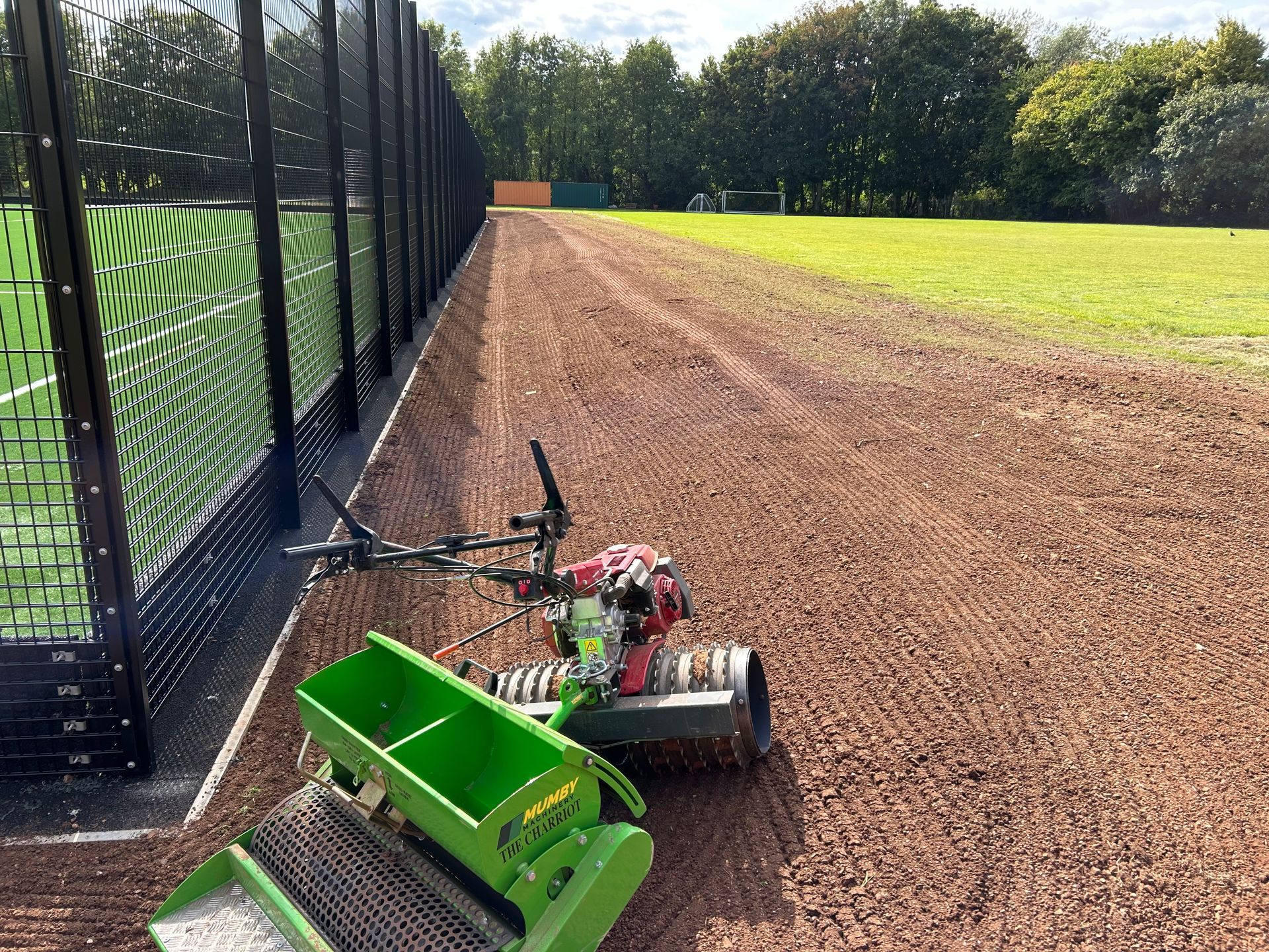 A green machine seeding a reddish-brown area next to a black fence and green field.