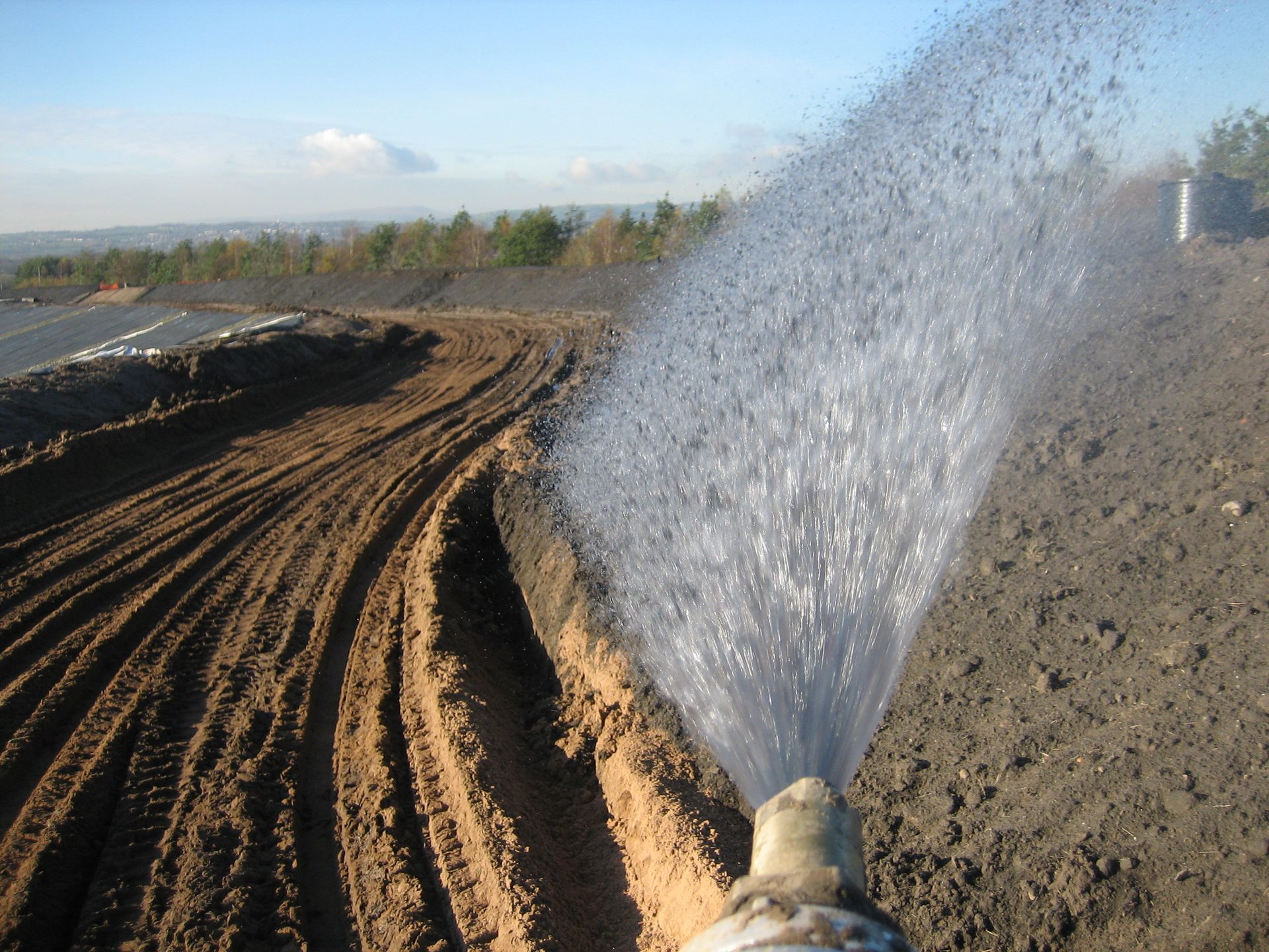 Water spraying onto brown dirt, possibly for dust control, in an outdoor area with a forest backdrop.