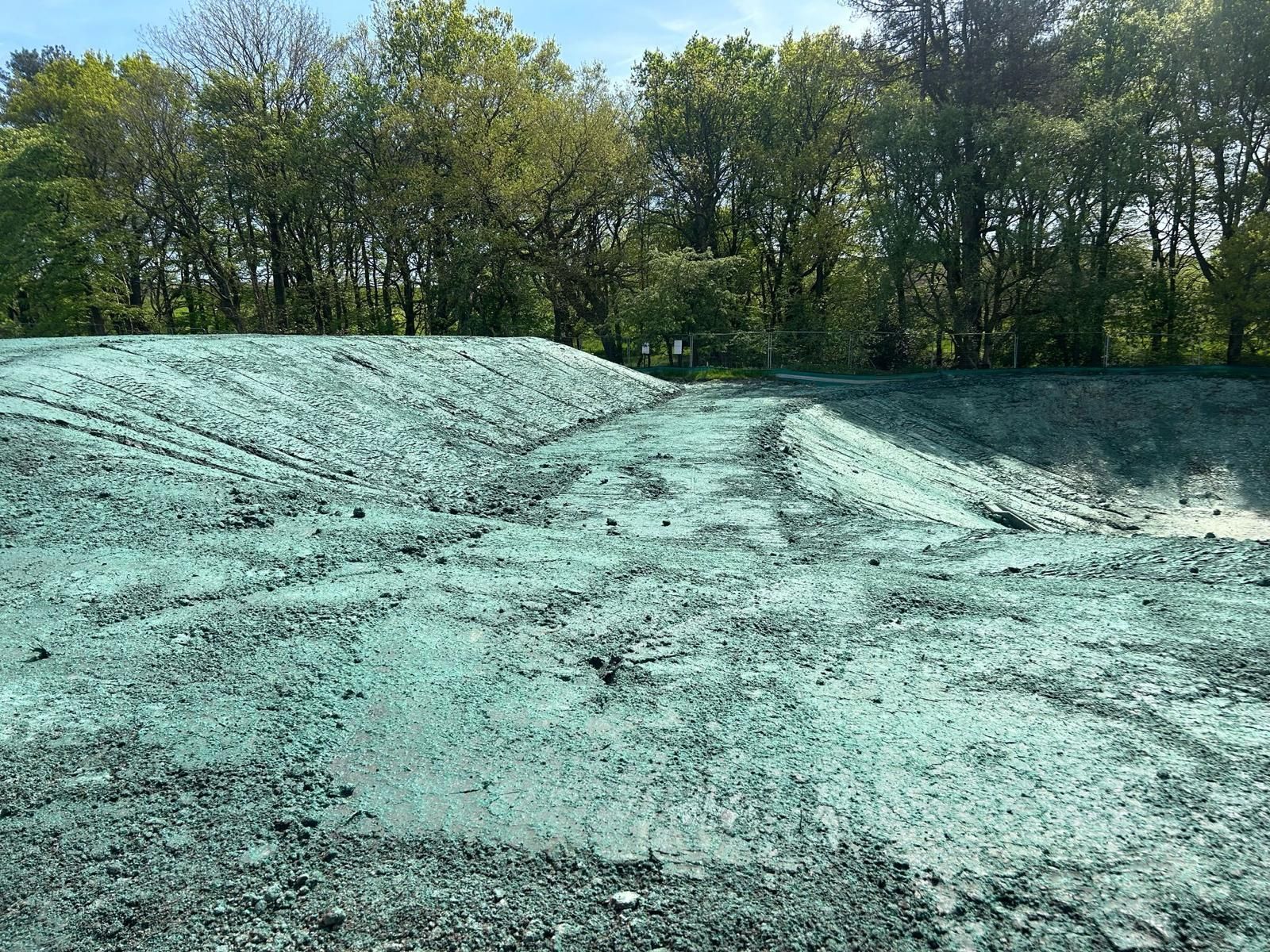 Green-covered dirt hill with tire tracks, trees in the background under a blue sky.