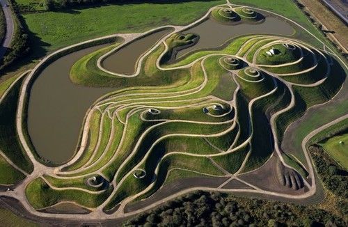 Aerial view of a green earthwork sculpture resembling a reclining woman. Features terraced hills, water, and pathways.