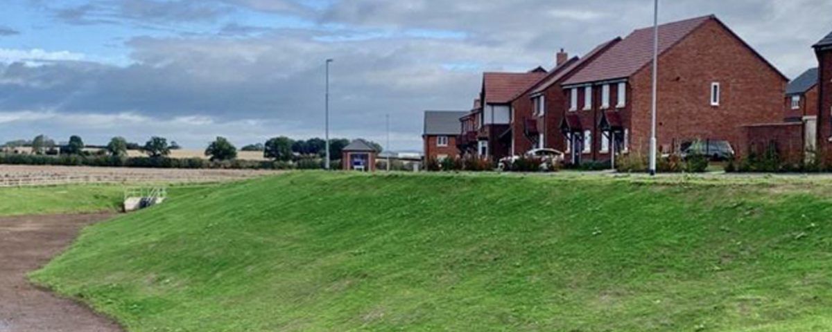 A green grassy hill leads to brick houses under a cloudy sky.