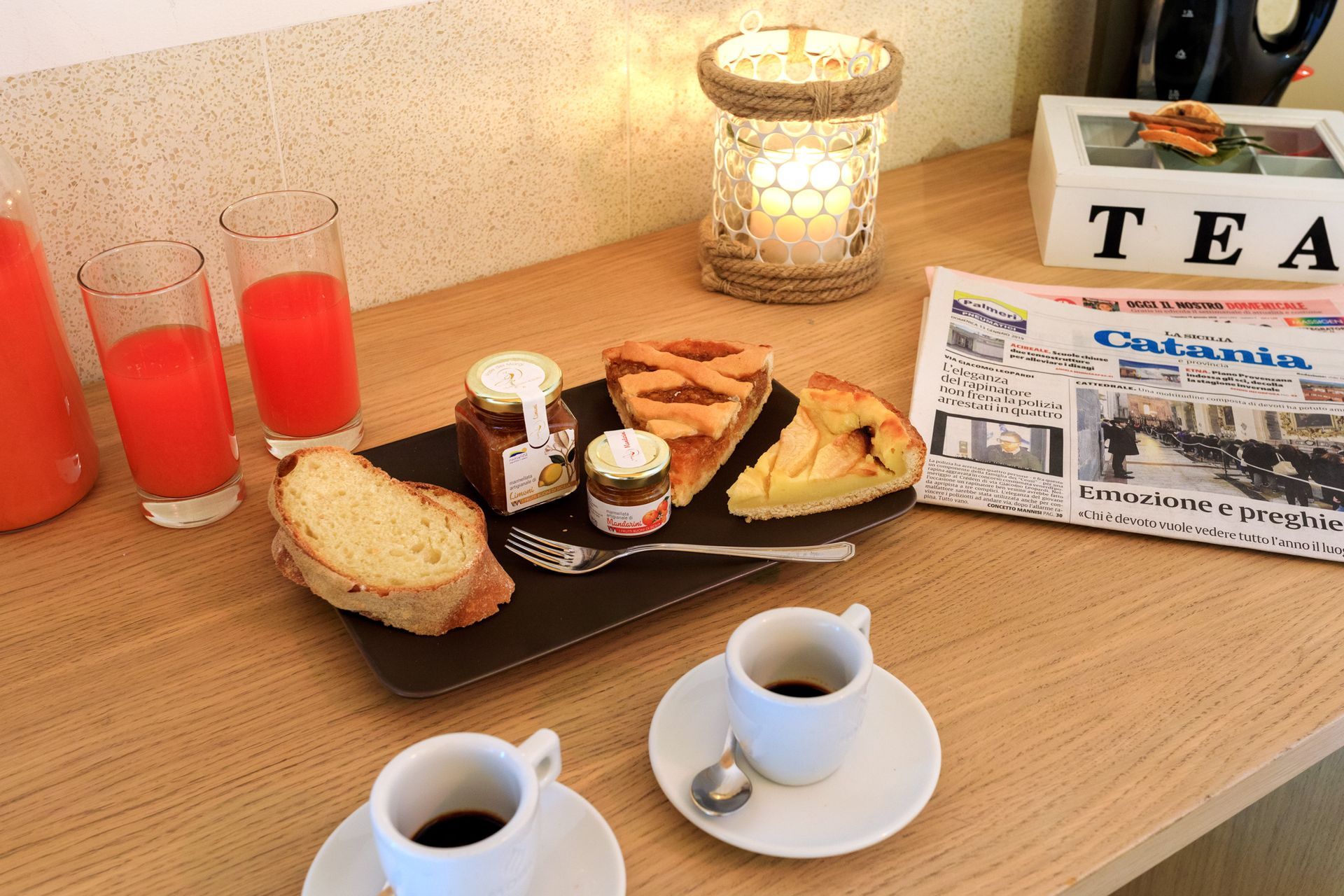 Tavolino da tè con pasticcini, due tazze di caffè, succo d'arancia, una candela accesa e un giornale su un bancone di legno.