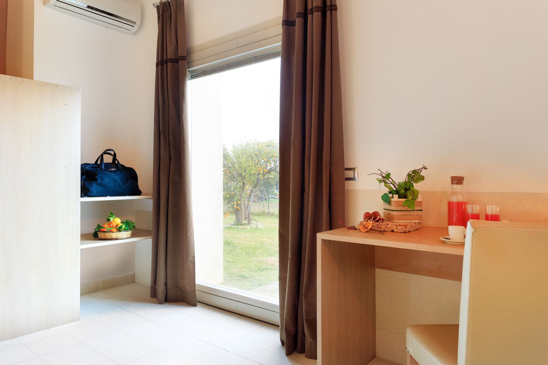 Una stanza luminosa con tende aperte, una grande finestra, mensole e una piccola scrivania in legno con piante e bottiglie.