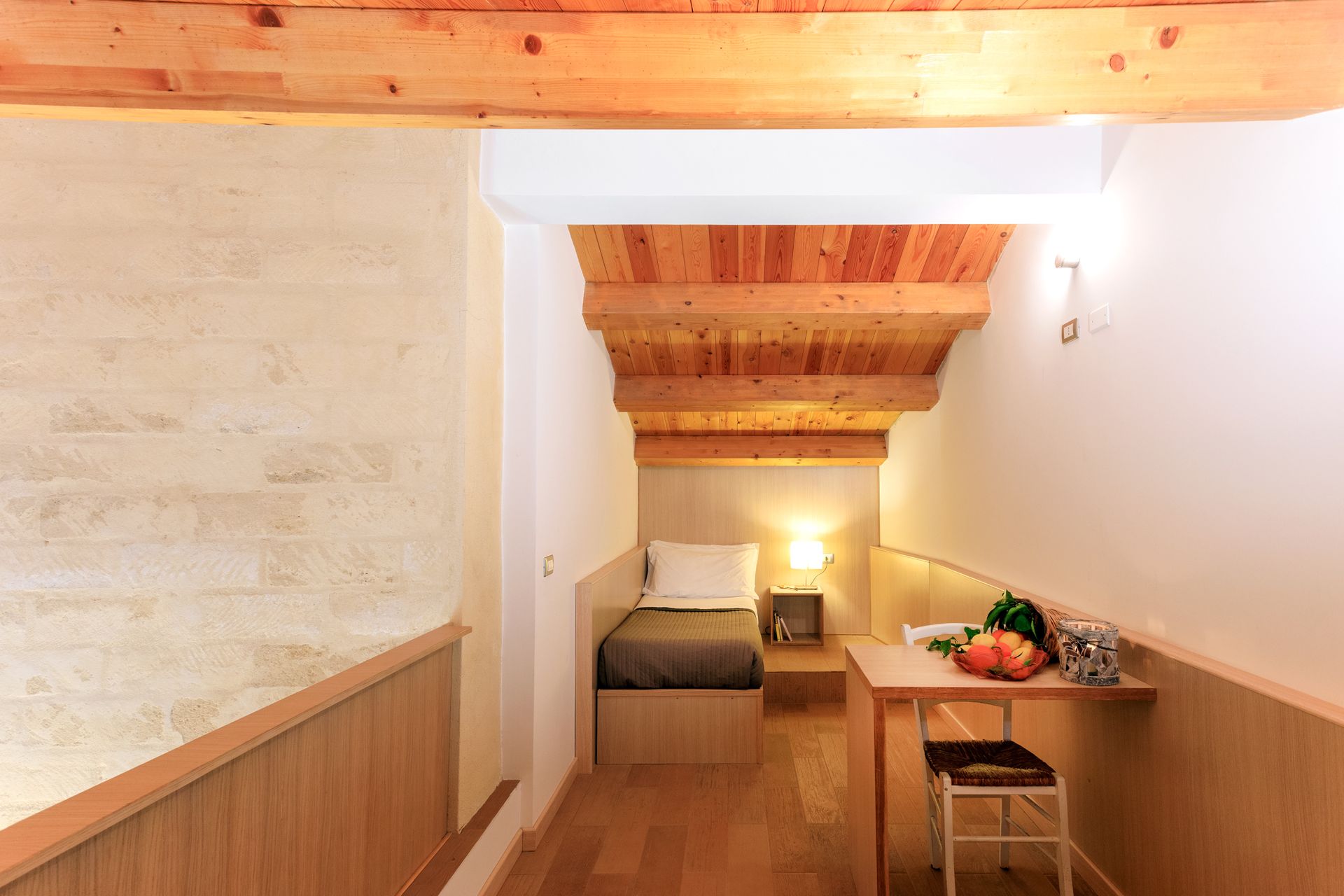 Camera da letto stretta in legno con letto, scrivania e luce calda sotto le travi in ​​legno a vista.