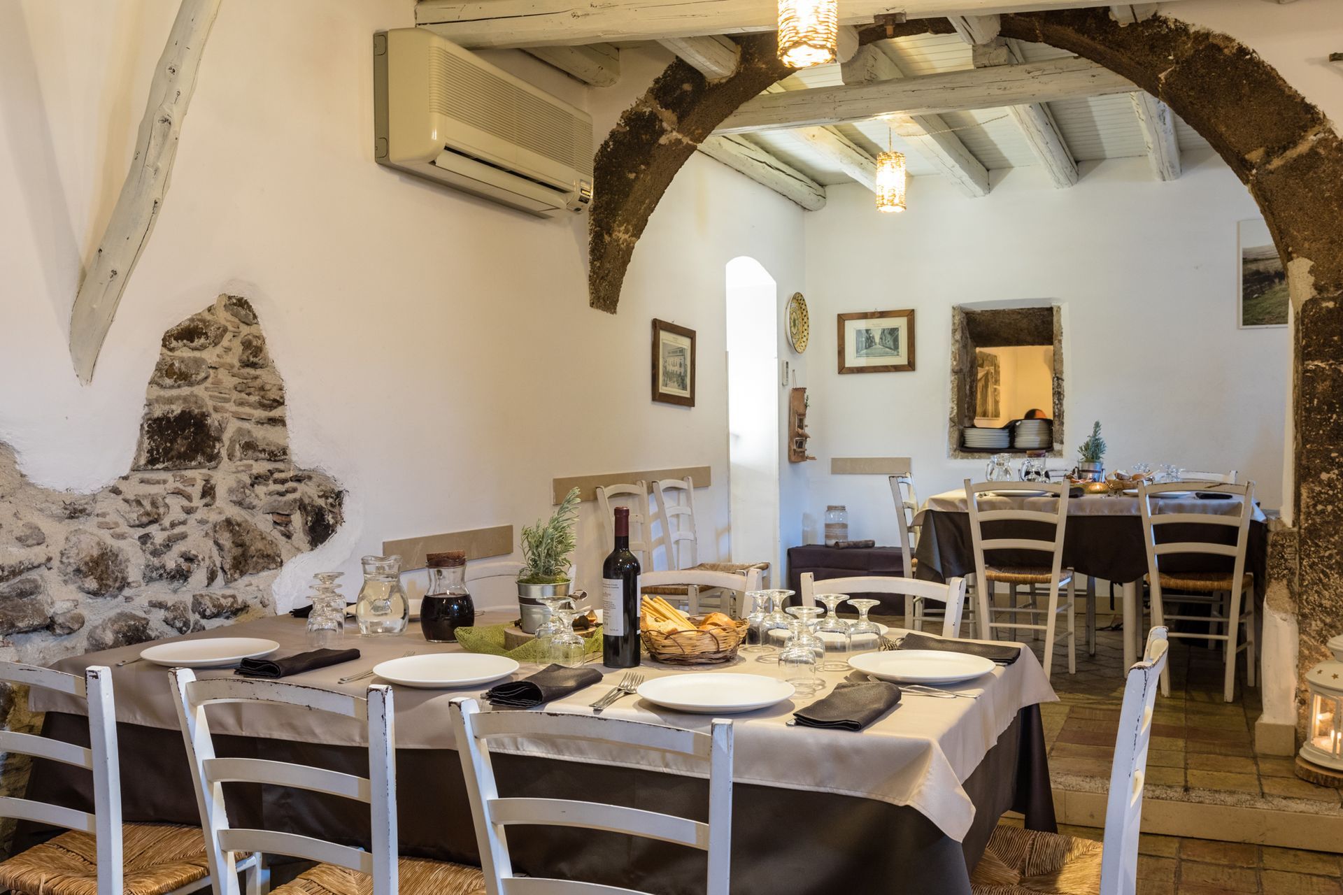 Sala da pranzo rustica di un ristorante con pareti in pietra, travi ad arco, sedie bianche e tavoli apparecchiati.
