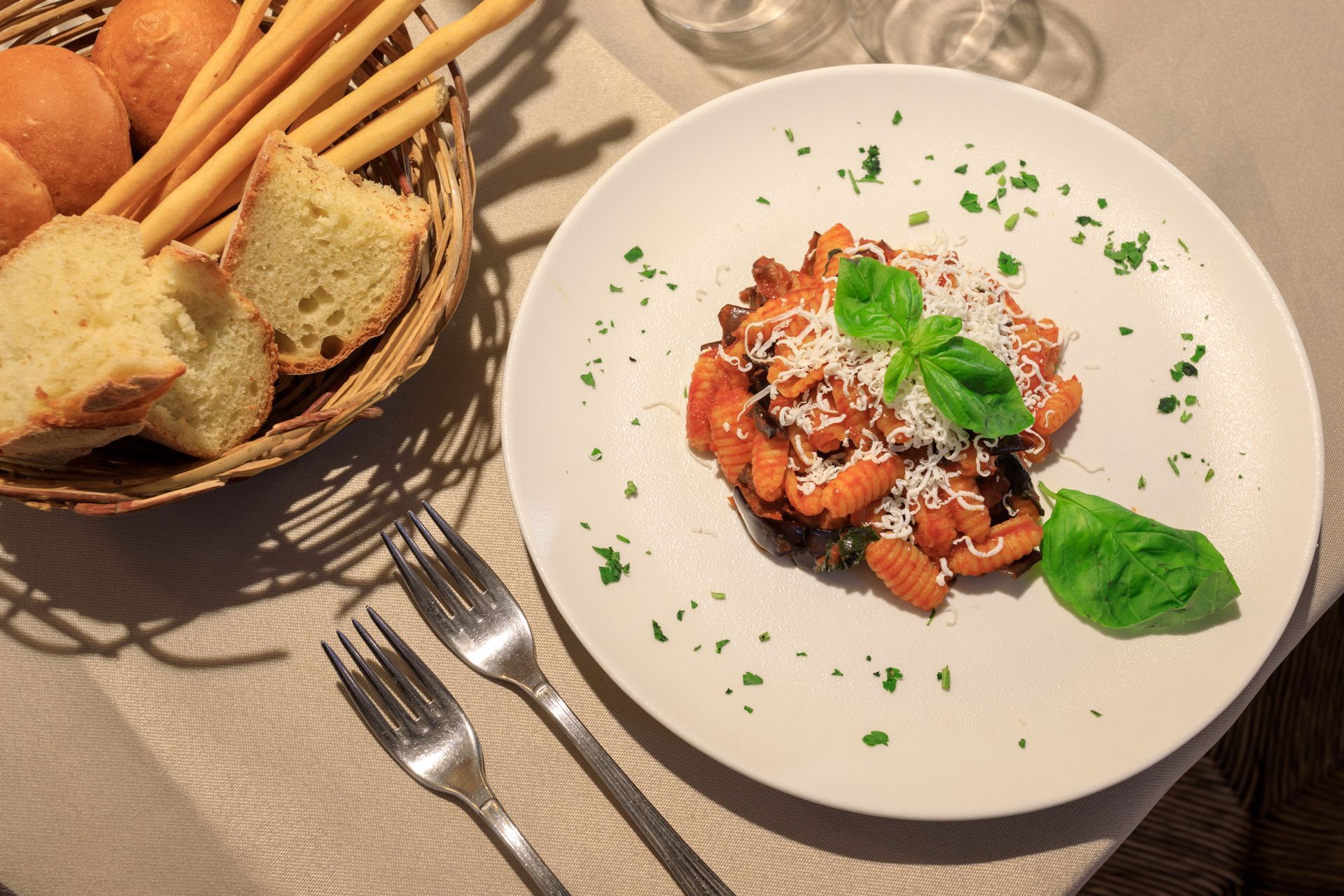 Un piatto di pasta con basilico e formaggio grattugiato accanto a un cestino di pane su un tavolo illuminato dal sole.