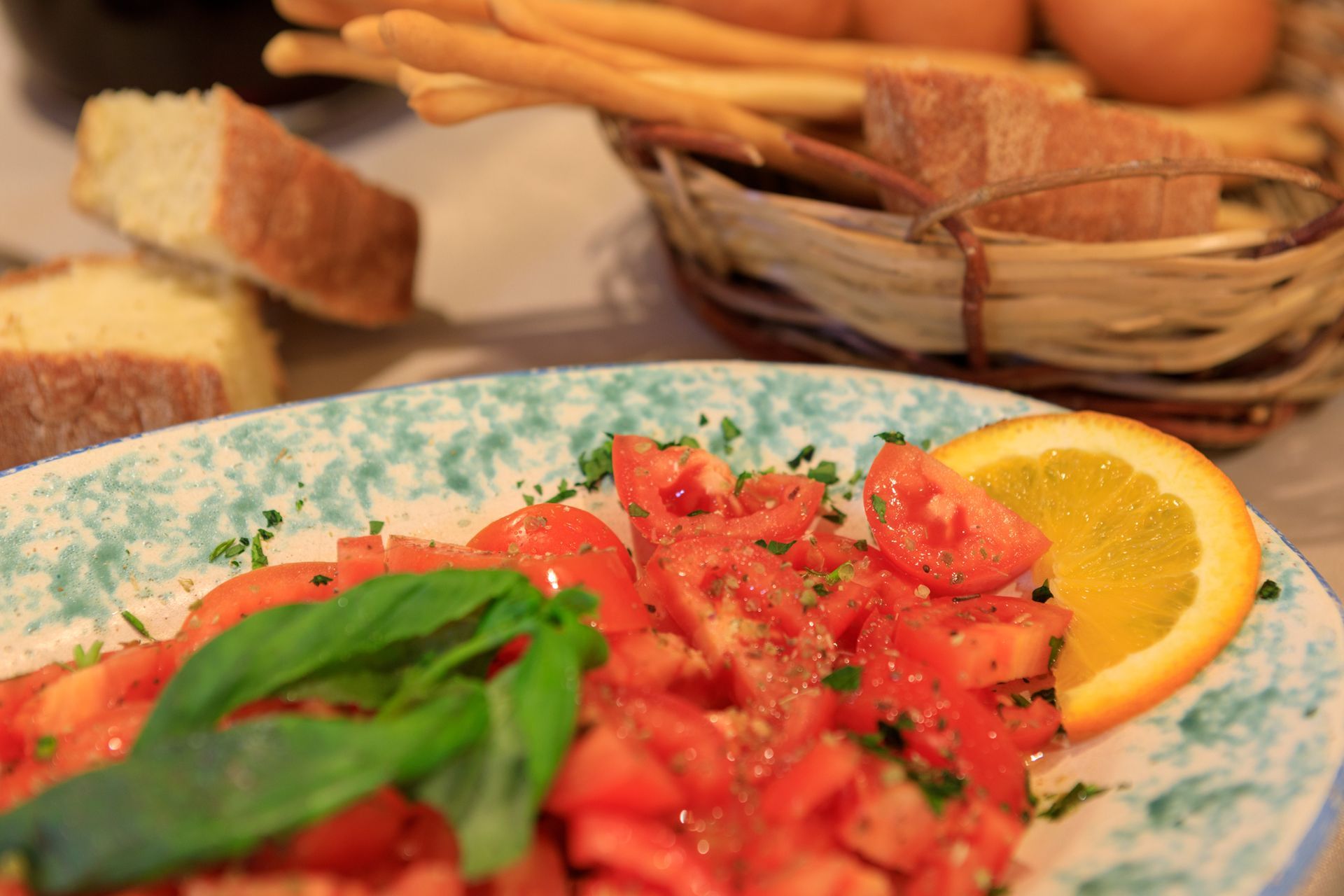 Insalata fresca di pomodori con basilico e limone su un piatto decorato, con pane e grissini sullo sfondo.