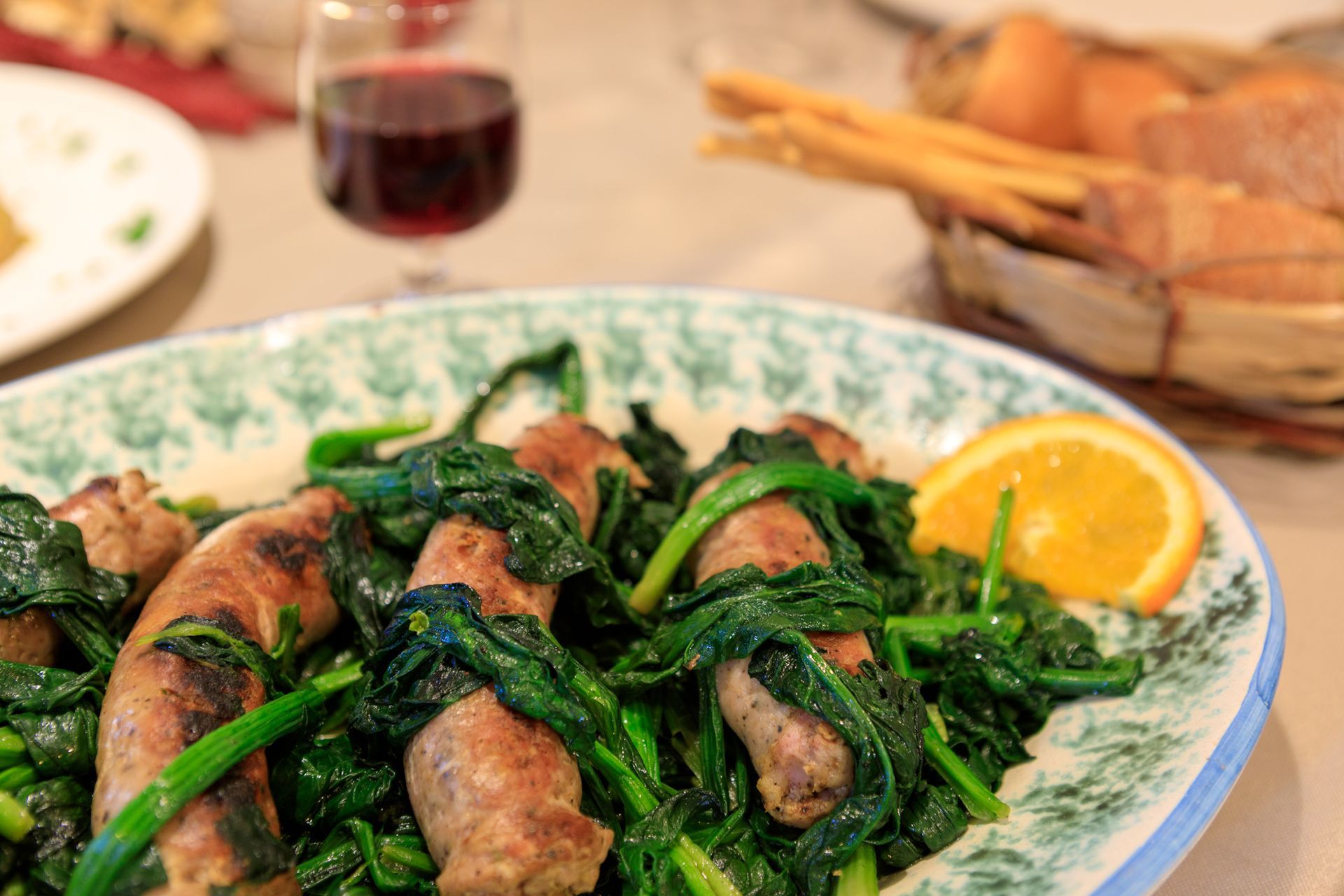 Un piatto di verdure e salsicce con uno spicchio di limone su un tavolo, con pane e vino sullo sfondo.