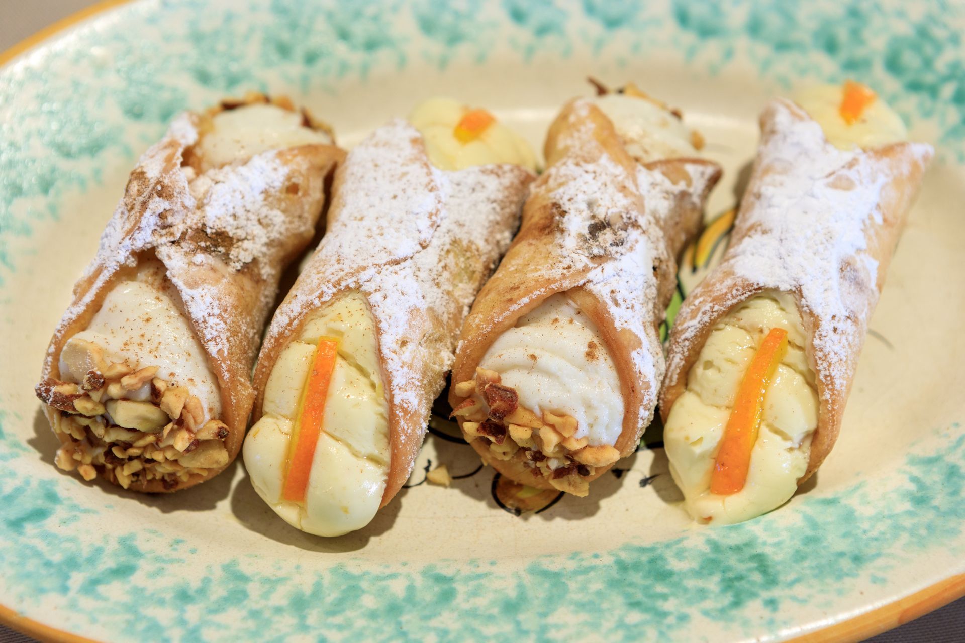 Quattro cannoli di zucchero a velo su un piatto decorativo, alcuni ripieni di pistacchi e scorza d'arancia.