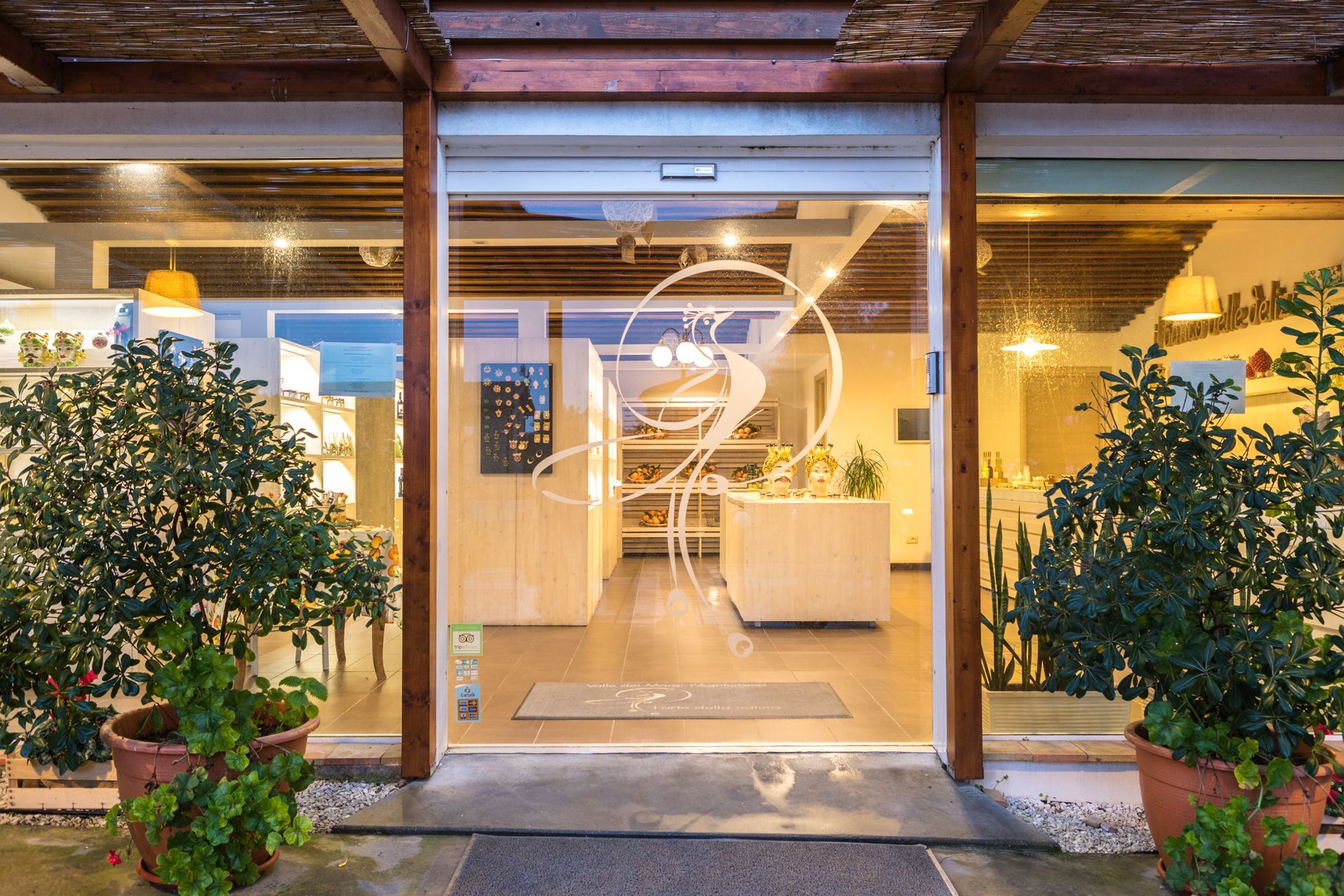 Vetrina con piante in vaso e ingresso interno illuminato da una luce calda.