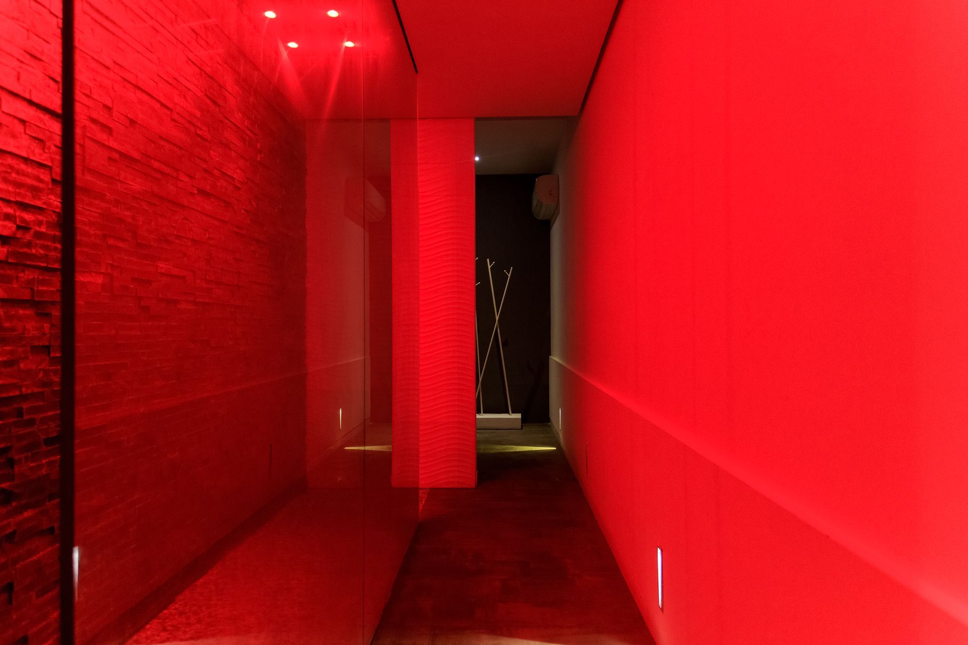 Stretto corridoio illuminato di rosso con un muro di mattoni sulla sinistra e una porta buia in fondo.
