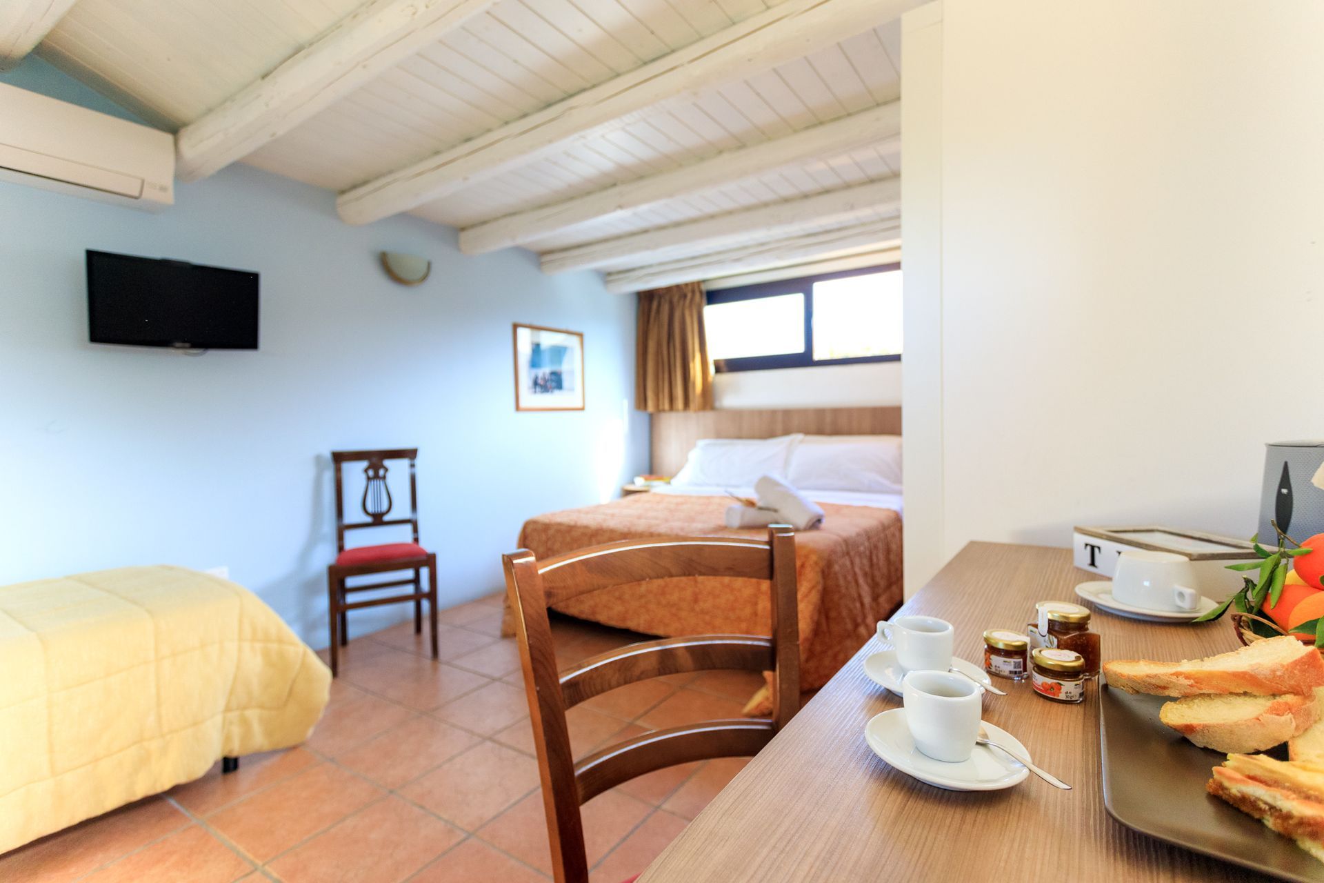 Piccola camera d'albergo con tre letti, scrivania, TV a parete e prodotti per la colazione vicino alla finestra.