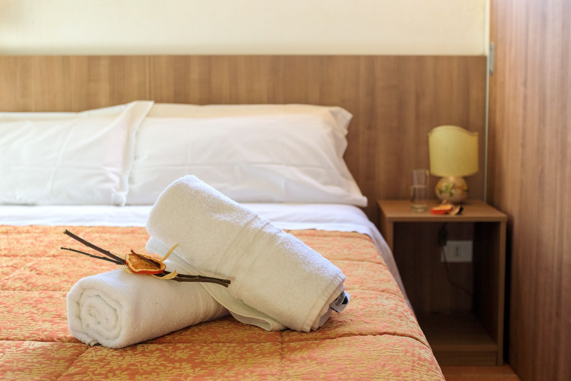 Letto d'albergo con asciugamani bianchi arrotolati e fiori su una coperta arancione, lampada da comodino a destra