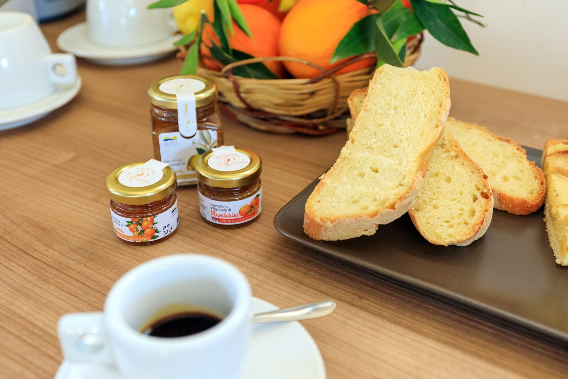 Caffè e pane tostato su un tavolo di legno con vasetti di marmellata e un cesto di frutta.
