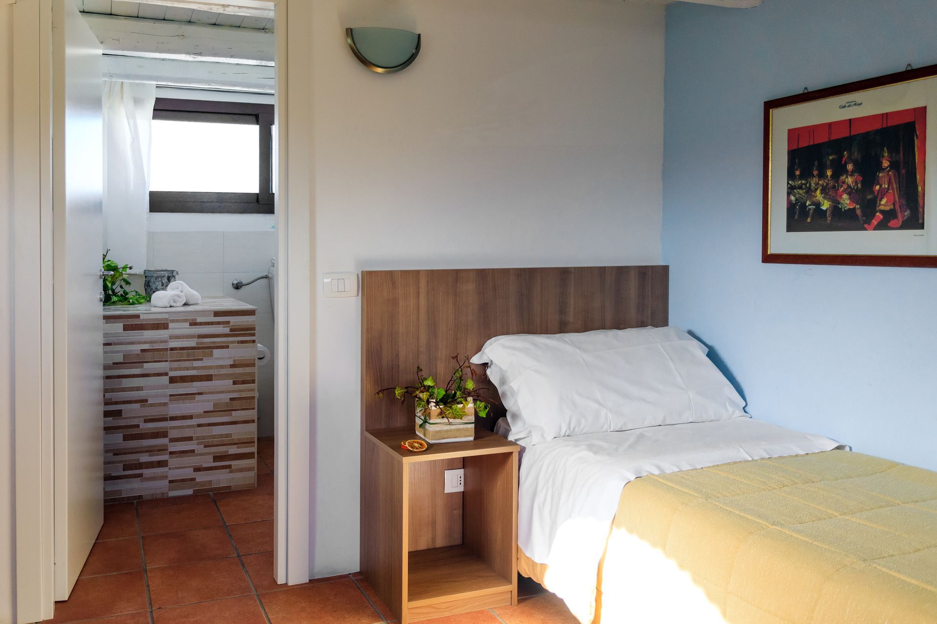 Piccola camera da letto con letto singolo, comodino in legno e porta che conduce a un bagno piastrellato.