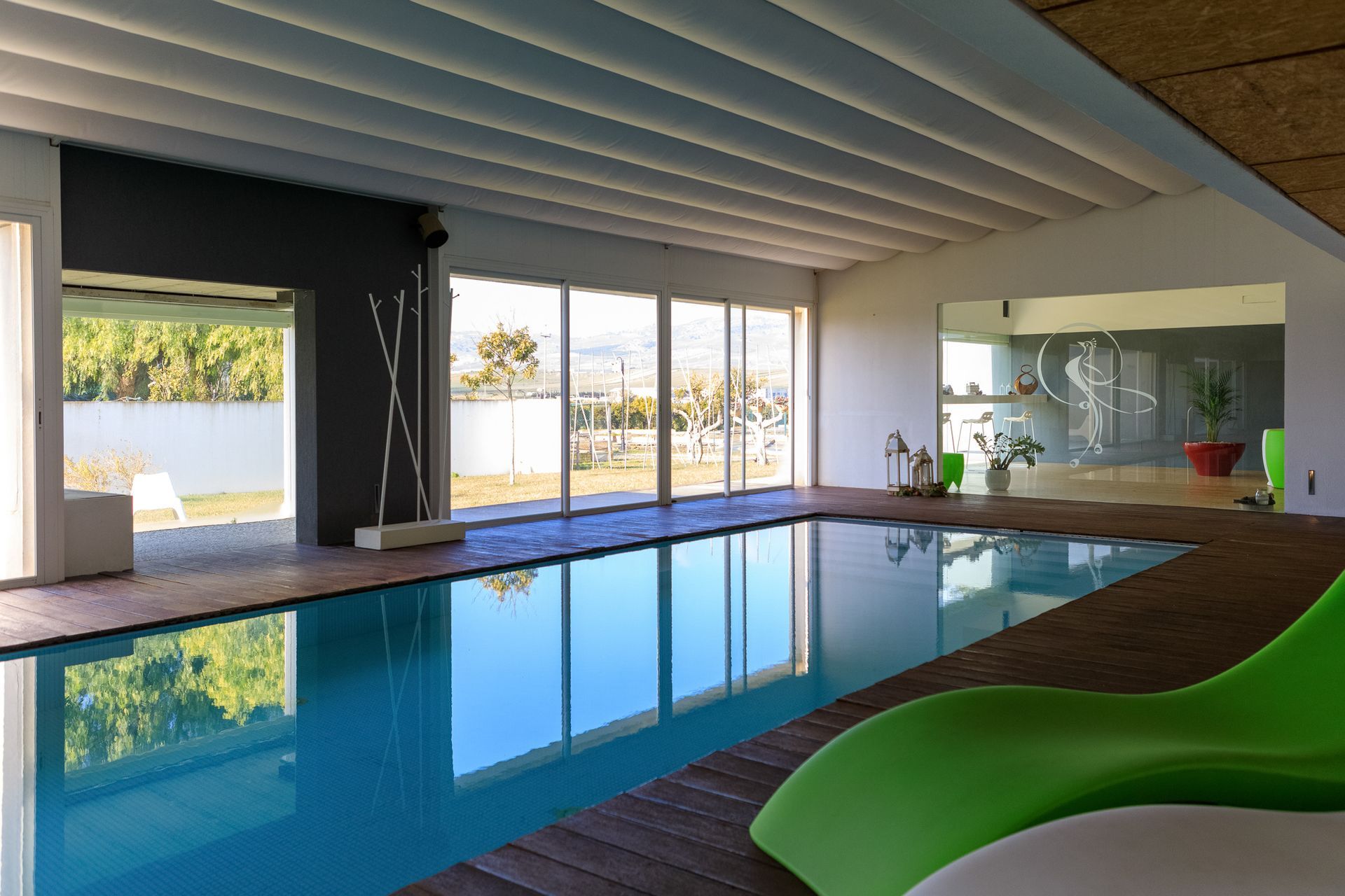 Piscina coperta con ampie vetrate, terrazza in legno e sdraio verde.