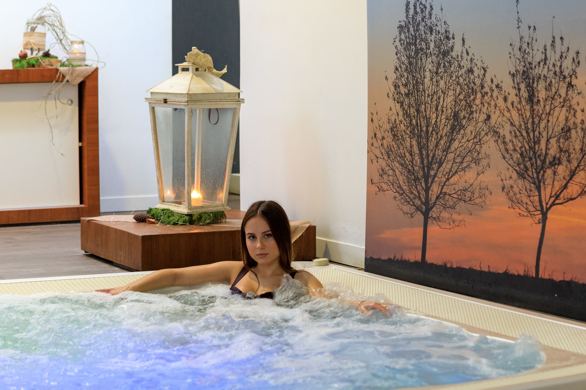 Una donna si rilassa in una vasca idromassaggio accanto a una lanterna accesa e a un quadro con un tramonto sullo sfondo.