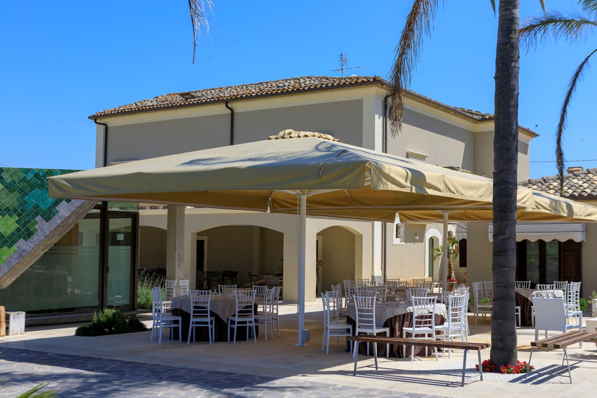 Dehor di un caffè all'aperto con ombrelloni beige e tavolini vuoti accanto a un edificio color crema sotto un cielo azzurro.