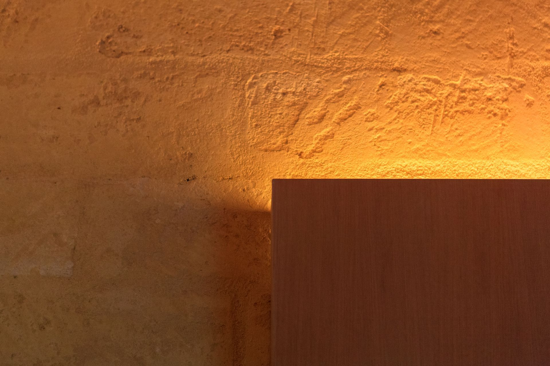 Una calda luce arancione illumina una parete ruvida accanto al bordo di un pannello di legno.
