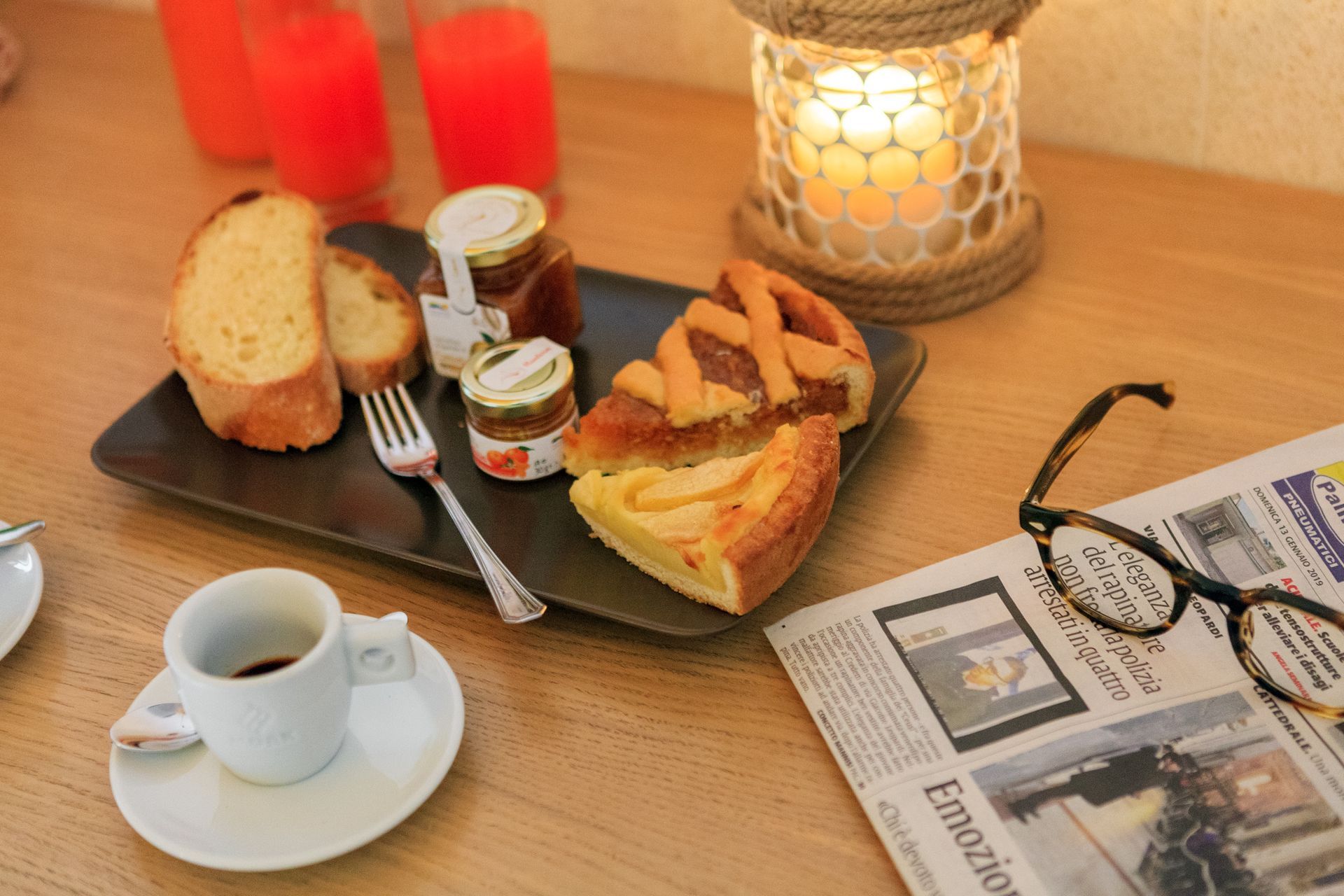 Tavolo da colazione illuminato da candele con caffè, pasticcini, marmellata, giornale e occhiali