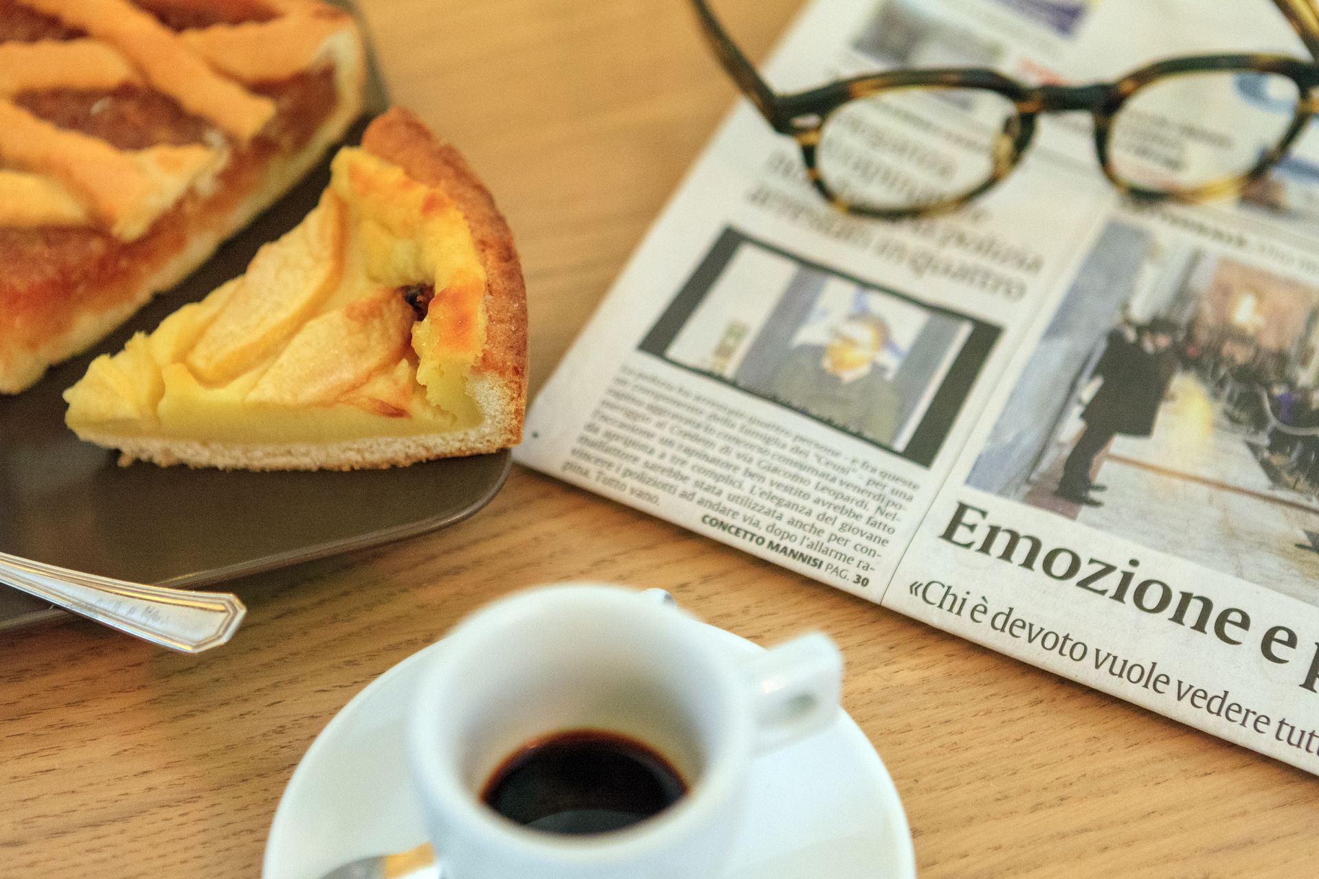 Una tazza di caffè accanto a un giornale aperto, occhiali e fette di pasticcino su un tavolo di legno.
