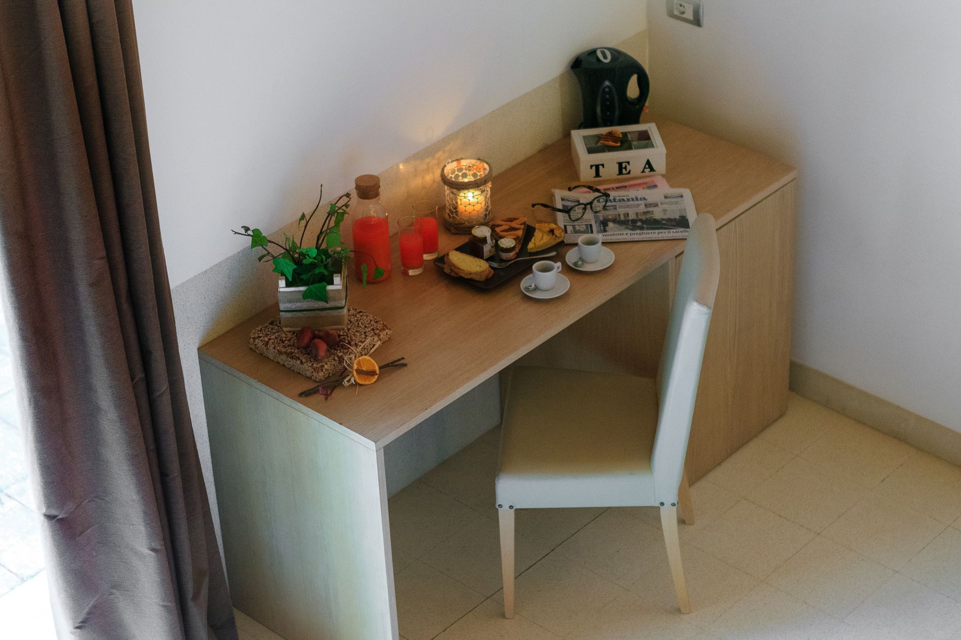 Un piccolo angolo studio con sedia, lampada, pianta e set da tè, sistemato vicino a una finestra.