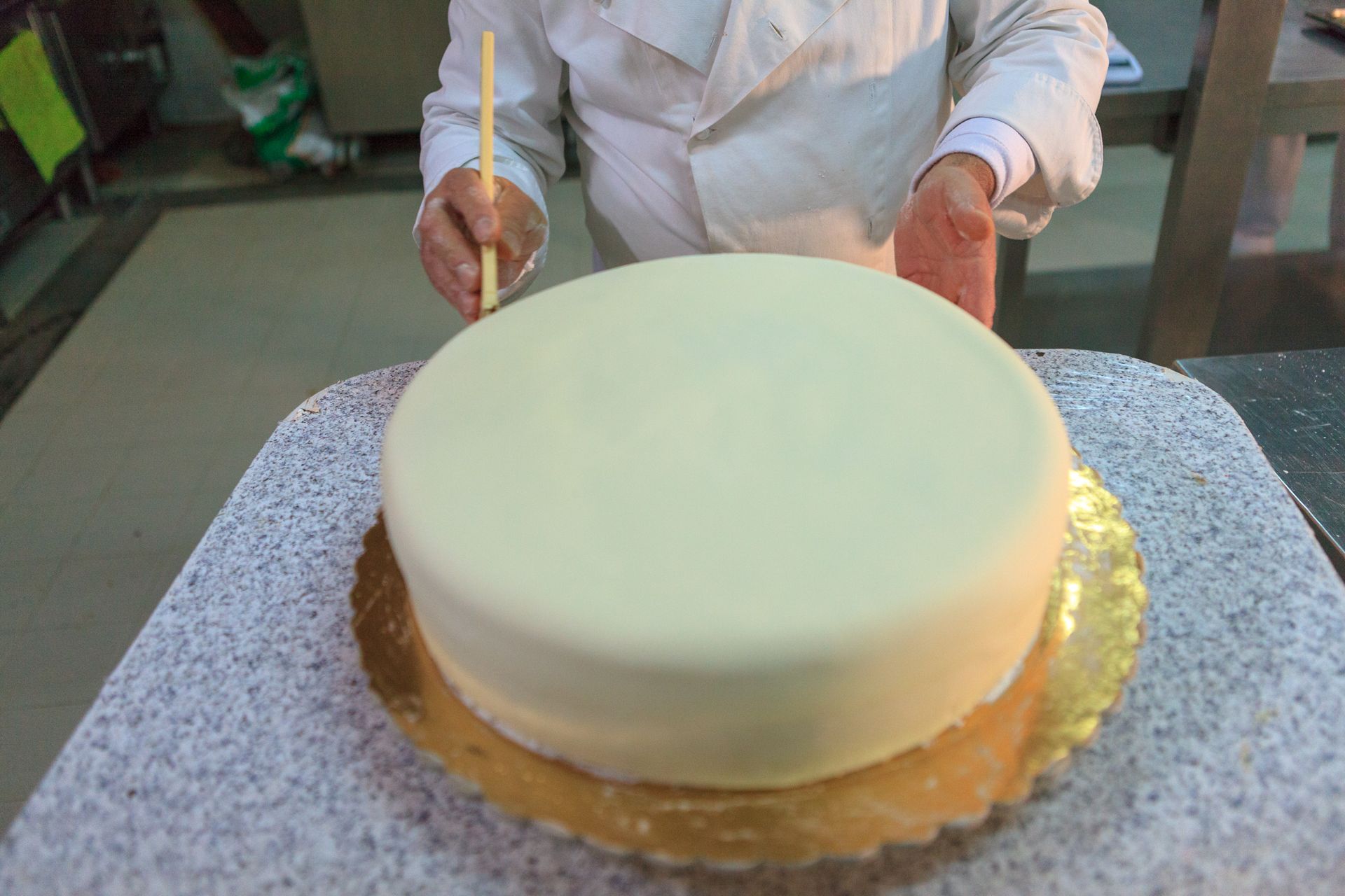 Una persona sta glassando una torta rotonda bianca su un piatto girevole di metallo in una cucina.