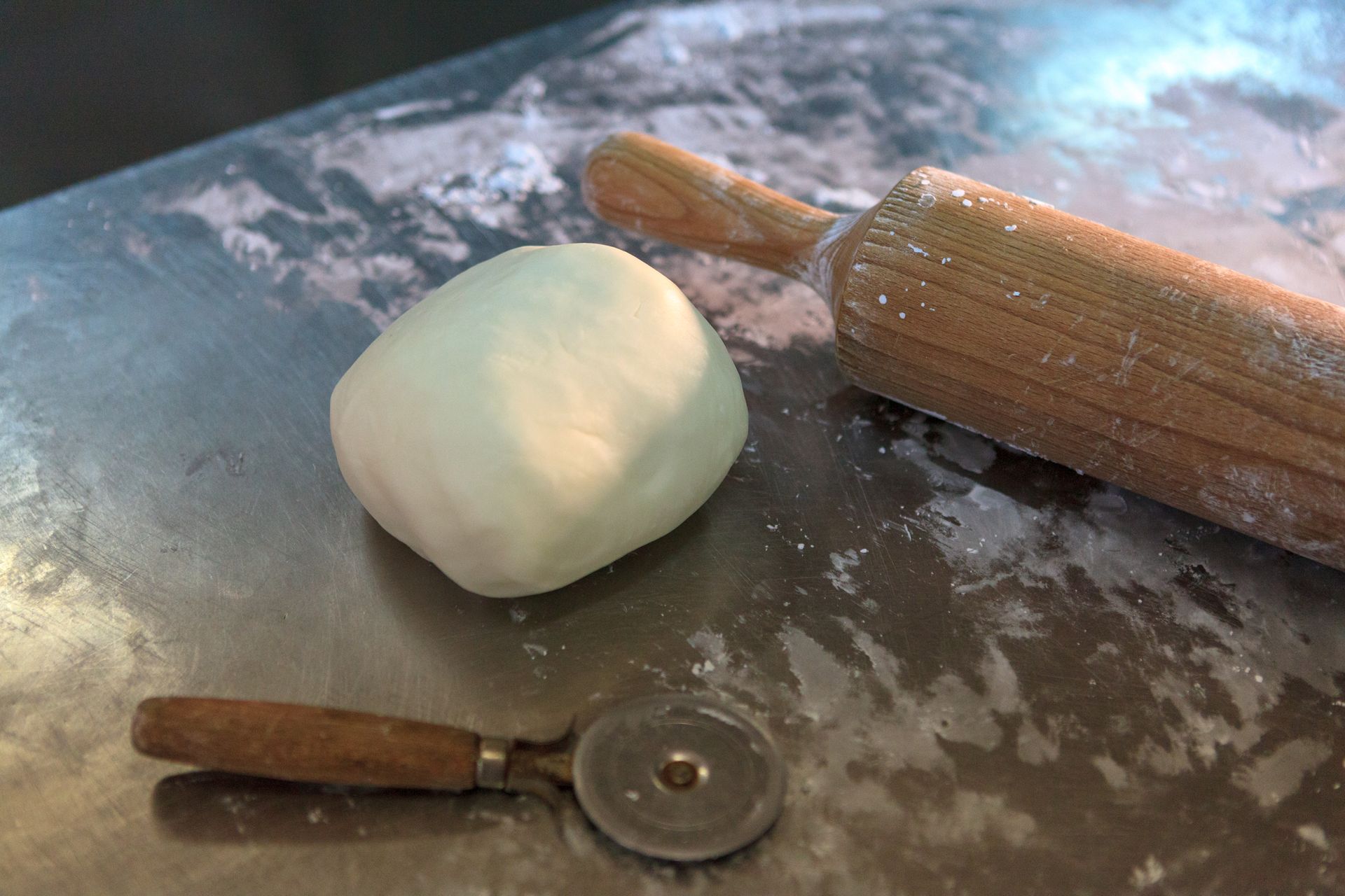 Una palla di impasto su un piano di lavoro infarinato, con un mattarello e una rotella per pasta nelle vicinanze.