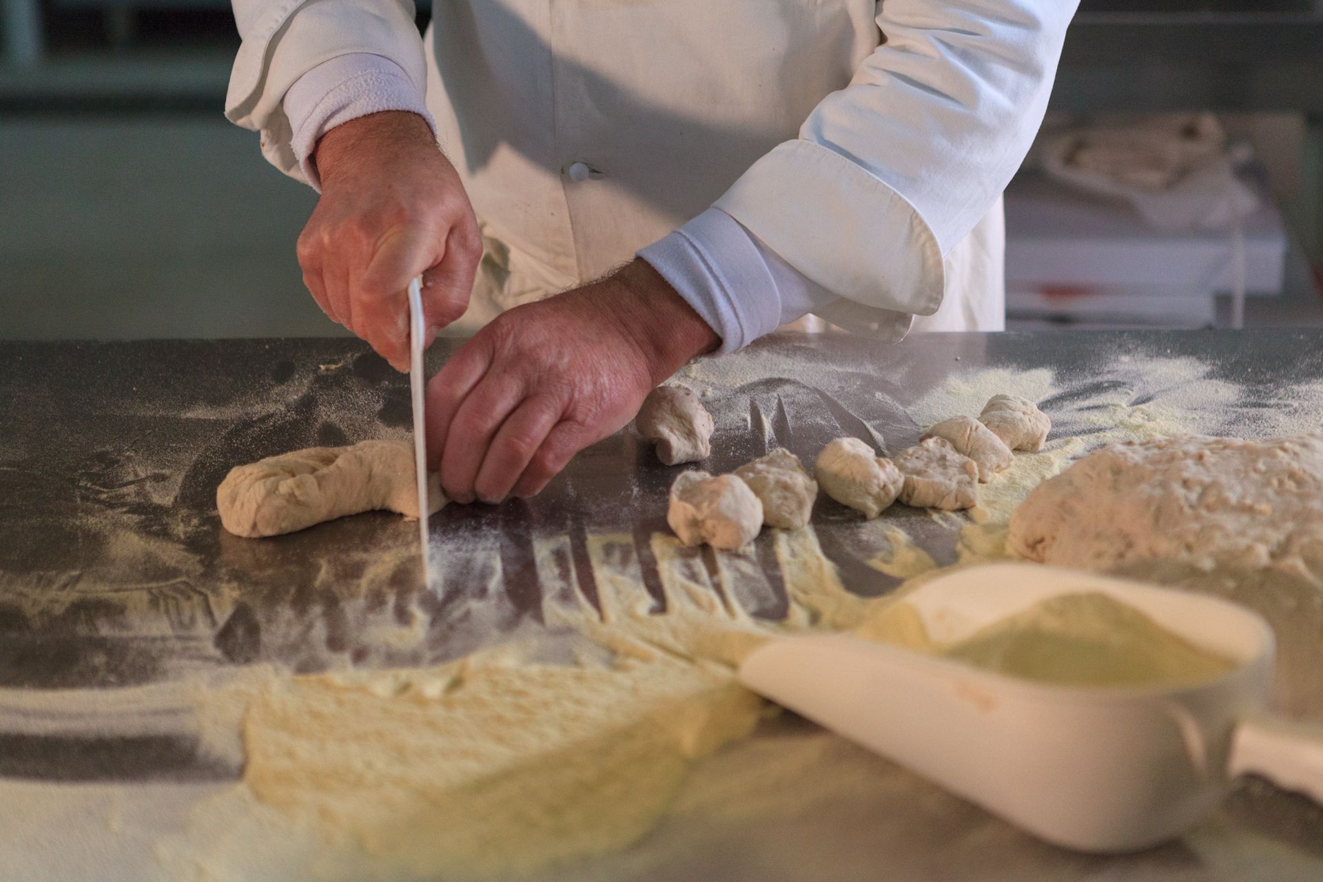 Mani che tagliano l'impasto su un piano di lavoro infarinato in una cucina, con ciotole di farina nelle vicinanze.
